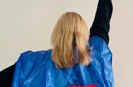 A woman wearing a superhero cape holding one arm up in the air as though she is about to start flying